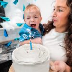 Mom holds a crying baby beside a white frosted cake with a single blue candle during a birthday celebration.