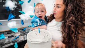 Mom holds a crying baby beside a white frosted cake with a single blue candle during a birthday celebration.
