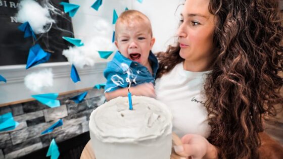 Mom holds a crying baby beside a white frosted cake with a single blue candle during a birthday celebration.