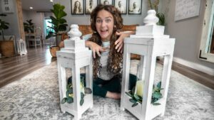 Girl sits cross-legged on a rug between two white wooden lanterns with candles and greenery, smiling excitedly.