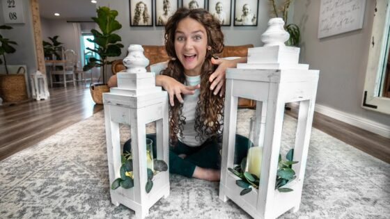 Girl sits cross-legged on a rug between two white wooden lanterns with candles and greenery, smiling excitedly.