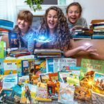 Three people (woman and two children) sit among a colorful pile of children's books on the living room floor, smiling at the camera.