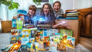 Three people (woman and two children) sit among a colorful pile of children's books on the living room floor, smiling at the camera.