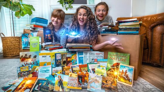 Three people (woman and two children) sit among a colorful pile of children's books on the living room floor, smiling at the camera.