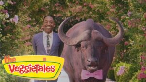 Smiling man in a blue suit stands beside a large buffalo wearing a pink bow tie, with a VeggieTales logo in the foreground and a floral backdrop.