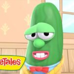 Green cucumber character from VeggieTales with droopy eyes and a red bow tie indoors.