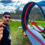 Man outdoors with a surprised expression holds a red dart toward a large looping metal slide and a small dartboard at the end.