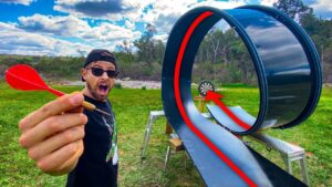 Man outdoors with a surprised expression holds a red dart toward a large looping metal slide and a small dartboard at the end.