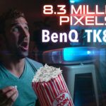 Man in a teal shirt looks surprised while watching a blue-lit BenQ TK850i projector, holding popcorn in his hand.