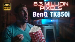 Man in a teal shirt looks surprised while watching a blue-lit BenQ TK850i projector, holding popcorn in his hand.