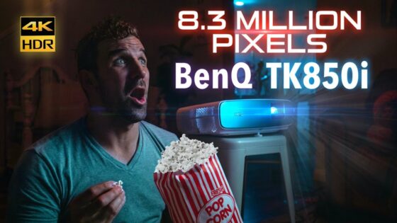 Man in a teal shirt looks surprised while watching a blue-lit BenQ TK850i projector, holding popcorn in his hand.