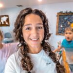 Smiling woman with braided hair posing with several children in a bright classroom filled with world maps and a chalkboard behind them.