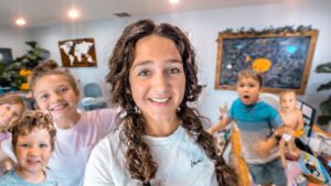 Smiling woman with braided hair posing with several children in a bright classroom filled with world maps and a chalkboard behind them.