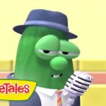 Green VeggieTales character in a fedora, singing into a retro microphone on a yellow stage with a colorful backdrop.