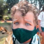 Child wearing a dark face mask at a zoo, close-up foreground with lions resting in the background.