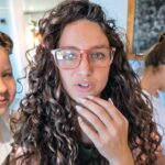 Teacher with curly hair and pink glasses speaks to the camera as children study at a table in a bright classroom.