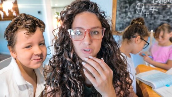 Teacher with curly hair and pink glasses speaks to the camera as children study at a table in a bright classroom.