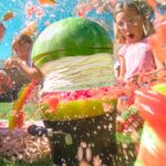 Group of kids outside having a water fight; a large watermelon prop sits on a black container while pink water splashes everywhere. - Informative image for a playful outdoor activity action scene.