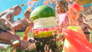 Group of kids outside having a water fight; a large watermelon prop sits on a black container while pink water splashes everywhere. - Informative image for a playful outdoor activity action scene.