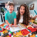 A woman and a child with curly hair playfully pose over a cluttered table of colorful building blocks, rulers, and activity books in a bright home.