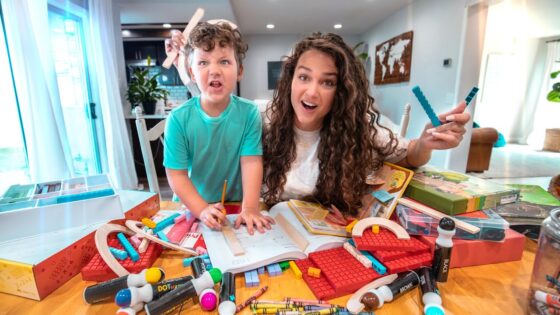 A woman and a child with curly hair playfully pose over a cluttered table of colorful building blocks, rulers, and activity books in a bright home.