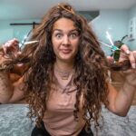 Woman sitting on the floor, holding scissors in each hand and teasing her long curly hair with a playful expression in a living room.