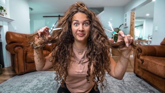 Woman sitting on the floor, holding scissors in each hand and teasing her long curly hair with a playful expression in a living room.