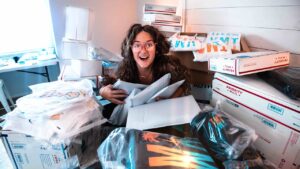 Young woman with curly hair and glasses surrounded by stacked mailboxes and shipping boxes, smiling while hugging padded envelopes.