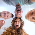 Group of four friends leaning over the camera, smiling and laughing as they look down at the lens from above.