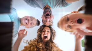 Group of four friends leaning over the camera, smiling and laughing as they look down at the lens from above.