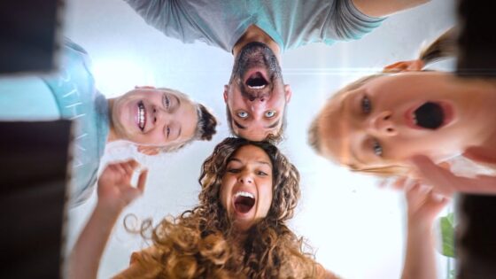 Group of four friends leaning over the camera, smiling and laughing as they look down at the lens from above.