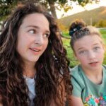 Two friends sit on a park path at sunset, the left woman with long curly hair and the right with a top bun, both looking at the camera.