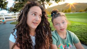 Two friends sit on a park path at sunset, the left woman with long curly hair and the right with a top bun, both looking at the camera.
