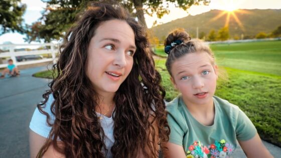 Two friends sit on a park path at sunset, the left woman with long curly hair and the right with a top bun, both looking at the camera.