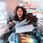 Young woman with curly hair and pink glasses sits amid stacked shipping boxes, hugging padded envelopes with a surprised smile.