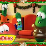 Three VeggieTales characters—Tomato, Broccoli with a scarf, and a green cucumber in a holiday sweater—sit on a couch in a festively decorated living room with a Christmas tree in the background.