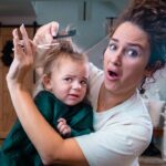 Mother trims daughter's hair at home while the child looks unhappy or uncertain during the haircut activity.