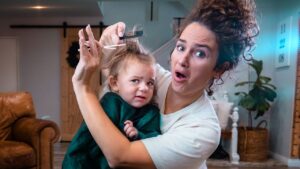 Mother trims daughter's hair at home while the child looks unhappy or uncertain during the haircut activity.