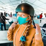 Front-facing girl in an orange hoodie wearing a teal face mask and helmet in a crowded indoor shelter or queue area