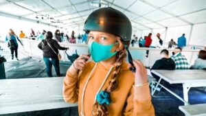 Front-facing girl in an orange hoodie wearing a teal face mask and helmet in a crowded indoor shelter or queue area