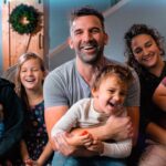 A smiling father in a gray t-shirt sits center, holding a laughing toddler while surrounded by siblings and a partner in a cozy living room.