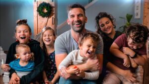 A smiling father in a gray t-shirt sits center, holding a laughing toddler while surrounded by siblings and a partner in a cozy living room.