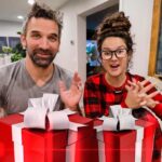 Two people smile at the camera as they present two large red gift boxes with white ribbons on a table, holiday scene ahead.
