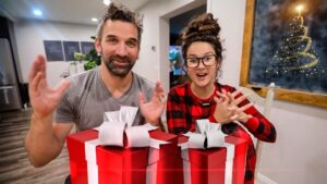 Two people smile at the camera as they present two large red gift boxes with white ribbons on a table, holiday scene ahead.
