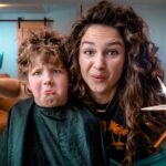 Woman cutting a child's hair at home, holding a pair of scissors and a blow dryer while the child sits in a cape in a cozy living room