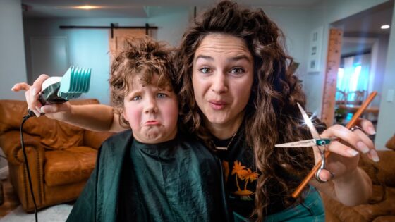 Woman cutting a child's hair at home, holding a pair of scissors and a blow dryer while the child sits in a cape in a cozy living room