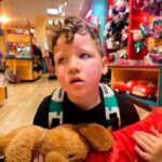Young boy in a toy store holding a brown teddy bear and a red plush dragon, looking away with a backpack on.