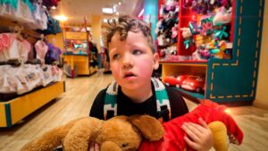 Young boy in a toy store holding a brown teddy bear and a red plush dragon, looking away with a backpack on.