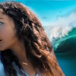 Woman in a white top, side profile looking to the left, with a large turquoise ocean wave behind her.
