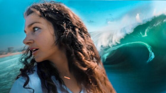 Woman in a white top, side profile looking to the left, with a large turquoise ocean wave behind her.